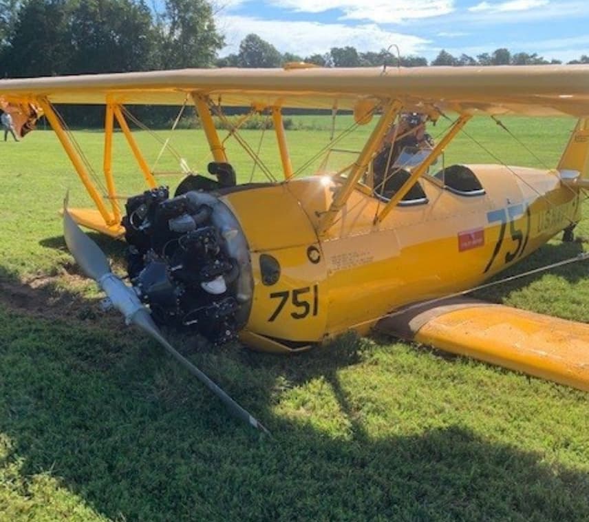 A yellow Boeing Stearman biplane sits damaged on a grassy field. The aircraft’s propeller is bent, and the engine shows signs of impact. The number “751” is prominently painted on the nose and fuselage, and a pilot is still seated in the rear cockpit. The wings remain intact, and the surrounding area is bathed in clear daylight with trees in the background.